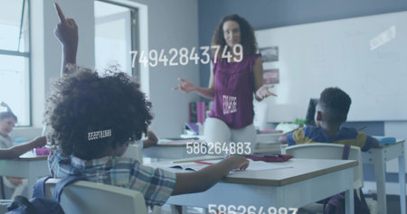 Raising hand curly-haired boy sitting at desk in classroom, with teacher by whiteboard and posters....