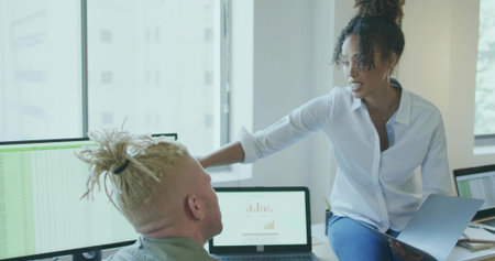 Discussing data woman in white shirt gesturing at man in office workspace, with monitors and laptop. Collaboration, teamwork, analytics, modernity, productivity, professional, digitalの写真素材