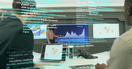 Presenting woman wearing business attire sharing charts in boardroom, with laptop and wall screens....