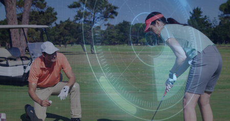 Kneeling golfer in orange polo placing ball on fairway, partner in green-polo swinging club by cart. Athletic, leisure, outdoor, precision, collaboration, scenic, sportの写真素材