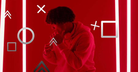 Standing man adjusting headphones in red-lit studio, wearing hoodie with neon strips and overlays. Urban, futuristic, vibrant, technology, entertainment, multimedia, innovationの写真素材