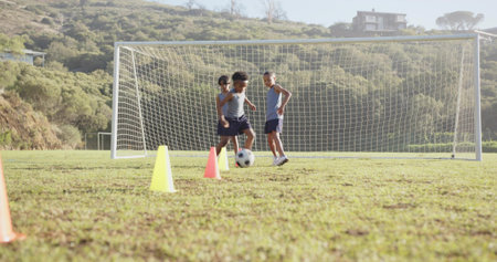 Three boys practicing soccer drills in grassy field, with ball and cones near goal. Athletics,...