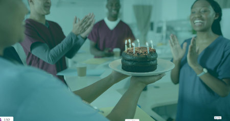 Presenting nurse holding two-tiered chocolate cake with lit candles in lounge with team clapping....