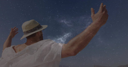 Man wearing loose white shirt stretching arms under Milky Way on plain, with straw hat. Astronomy,...