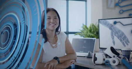 Smiling Asian female engineer sitting at desk in modern office with laptop and holographic overlay....