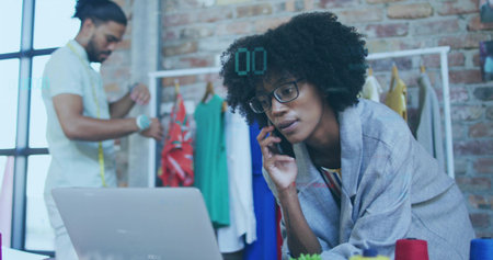 Female designer in shawl leaning over laptop, speaking on smartphone in studio with measuring tape. Creative workspace, fashion industry, tailoring, craftsmanship, collaborative, urban interiorの写真素材