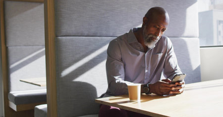 Holding smartphone, senior Black man wearing shirt sitting at modern lounge table with coffee cup. Contemporary, minimalist, nostalgic, professional, relaxed, natural, elegantの写真素材