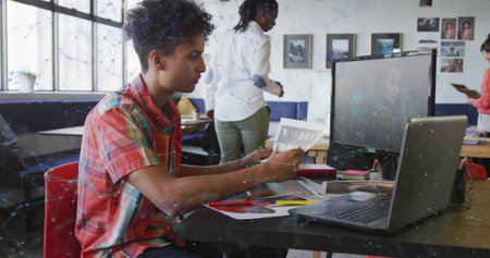 Reviewing printed proofs, man in red plaid shirt sitting at desk in design studio with laptop....