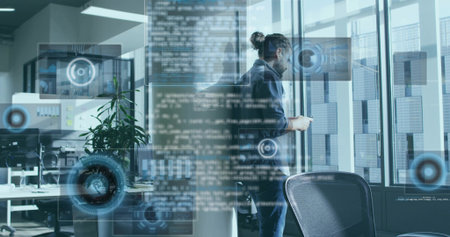 Holding smartphone hoodie-wearing man collaborating in office with dual monitors and HUD graphics. Technology, data, innovation, collaboration, modern, urban, analyticsの写真素材