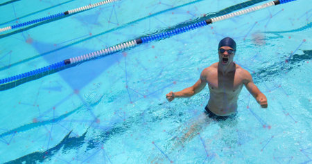 Emerging male swimmer raising fists from lane dividers in blue water, with swim cap goggles briefs....