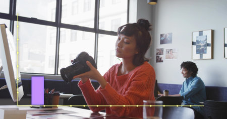 Inspecting DSLR camera Asian woman in red knitted sweater at modern shared workspace, with prints. Creative, collaborative, bright, professional, modern, inspiring, artisticの写真素材