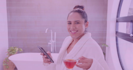 Smiling woman wearing white robe holding smartphone and tea mug in modern bathroom with tub, faucet. Minimalist, wellness, relaxation, skincare, luxury, natural light, sereneの写真素材