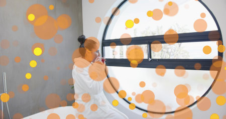 Holding white ceramic mug, Indian woman in bathrobe gazing through circular window in spa bathroom. Serenity, relaxation, wellness, minimalism, tranquility, interior, lifestyleの写真素材