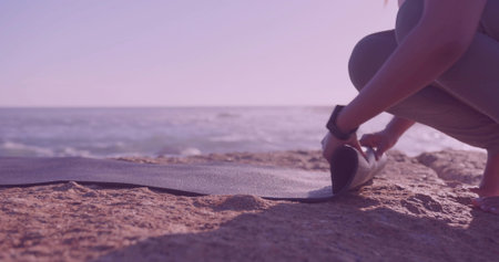 Unrolling Indian woman in leggings wearing smartwatch preparing mat on shore by ocean, copy space. Fitness, outdoor, activewear, wellness, solitude, nature, relaxationの写真素材