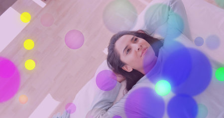 Reclining woman gazing upward on bed in bedroom at home, with pillow and abstract colored circles. Contemplative, relaxation, dreamy, abstract, tranquil, wellness, modernの写真素材