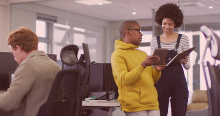 Standing man in yellow hoodie pointing at tablet in office, with coworker in overalls by monitors. Collaboration, teamwork, modern, professional, digital, creative, positiveの写真素材