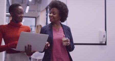 Walking coworkers in formal attire through office hallway, pointing at laptop, holding coffee cup. Professional, collaboration, corporate, communication, technology, modern, productivityの写真素材
