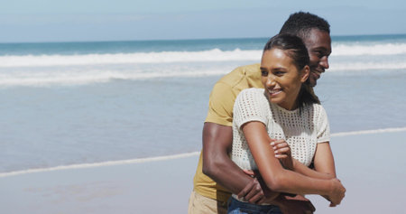 Hugging couple wearing casual summer clothes on sandy beach at water's edge, with ocean waves....