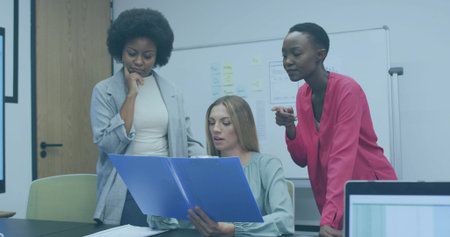 Reviewing notes on whiteboard, three women consulting laptop with blue folder in conference room....