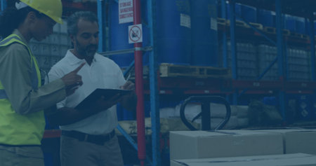 Checking stock, colleagues wearing hard hat and vest pointing at clipboard in warehouse, copy space....