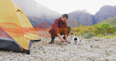 Crouching camper driving tent stake into rocky mountain plain, beside yellow tent, dog observing....