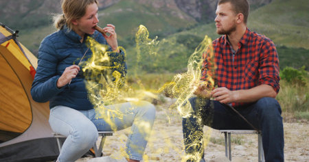 Roasting snack skewers camping couple sitting on folding stools at mountain campsite, yellow tent. Adventure, outdoor, rustic, cozy, exploration, wilderness, relaxationの写真素材