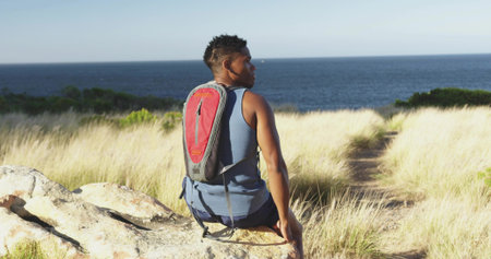Man wearing blue tank top sitting, gazing at ocean on dune rock. Adventure, resilience, exploration,...