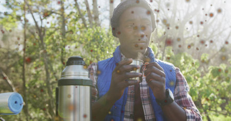 Man wearing blue vest sipping from metal cup in forest clearing, with thermos and sleeping pad. Adventure, solitude, wilderness, hydration, nature, exploration, rusticの写真素材