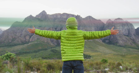 Standing hiker stretching arms wide facing away in foothills, wearing green jacket and blue jeans....