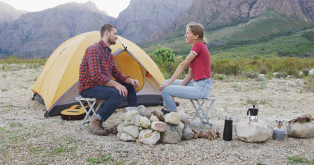 Sitting couple wearing flannel and red shirts camping at foothill, with yellow dome tent, fire pit....