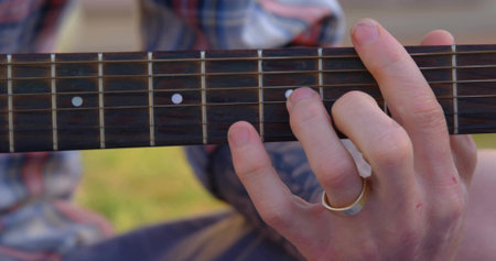 Pressing strings down on acoustic guitar fretboard in grassy park, showing plaid shirt, silver ring. Musician, craftsmanship, rustic, leisure, outdoor, harmony, detailの写真素材