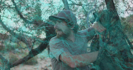 Climbing man wearing baseball cap and green t-shirt in dry scrubland, with tree branches and strap....