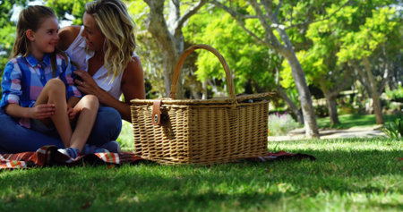 Sitting mother and daughter talking on plaid blanket in park with wicker picnic basket, copy space. Families, bonding, leisure, outdoor, serenity, connection, naturalの写真素材