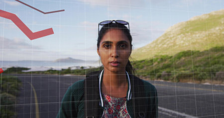Standing Indian woman wearing bomber jacket on paved coastal road, with backpack and sunglasses....