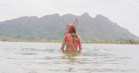Wading woman wearing swimwear waist-deep in lake water at mountain foothill, with rocky shoreline....