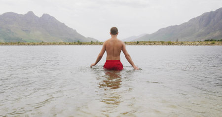 Touching water bare-chested man wading in lake, with red swim trunks and mountains. Solitude, serenity, outdoors, reflection, nature, landscape, minimalistの写真素材