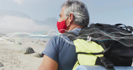 Standing man wearing red mask, blue shirt on beach with backpack and sleeping mat, copy space. Adventure, exploration, wilderness, outdoor, resilience, tranquility, ruggedの写真素材