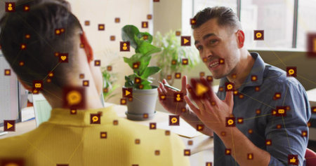 Gesturing man in blue shirt speaking at office desk near window, with plant, floating social icons. Collaboration, communication, professionalism, digital interaction, modernity, workspace, teamの写真素材
