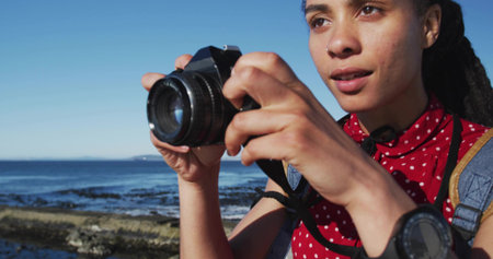 Shooting woman in red polka-dot shirt raising DSLR camera on rocky shore, with backpack, copy space. Adventure, exploration, maritime, leisure, nature, outdoor, scenicの写真素材