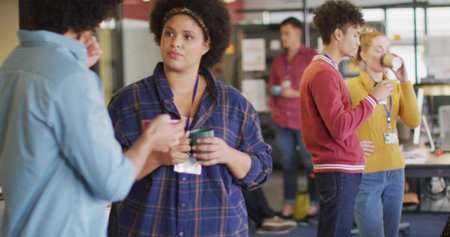 Chatting woman wearing blue plaid shirt holding green mug in office break area, with coffee mugs....