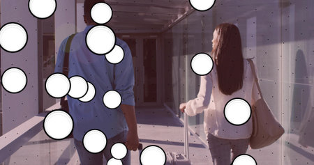 Walking couple rolling suitcases and carrying tote, crossbody bags in metal-framed glass corridor....