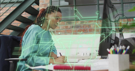 Writing woman wearing blue shirt focusing on notebook at desk in office, with hologram overlay. Creative, modern, innovative, professional, collaborative, technical, workstationの写真素材