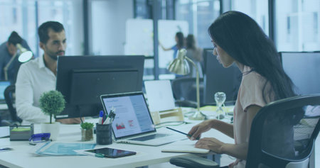 Reading documents, woman wearing blouse typing on laptop in open office, with notebook, whiteboards. Collaboration, teamwork, modernoffice, creativeworkspace, coworking, productivity, professionalの写真素材