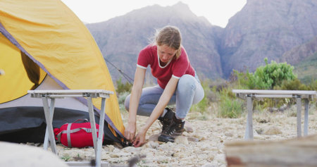 Crouching female camper securing yellow tent corner at rocky mountain campsite, with red duffel bag. Adventure, outdoor, exploration, rugged, nature, solitude, landscapeの写真素材