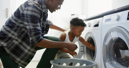 Helping parent and child loading laundry into front-loading washer in laundry room with gray basket. Domestic, family, caretaking, everyday, cozy, harmonious, interactionの写真素材