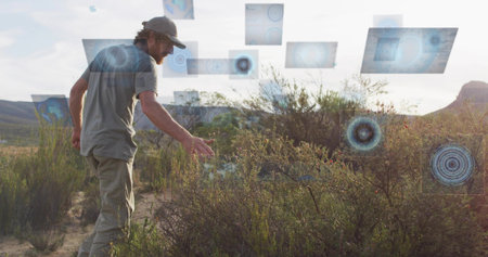 Analyzing man wearing baseball cap reaching toward shrubs with digital panels in desert, copy space. Technology, innovation, exploration, documentary, futuristic, professional, environmentalの写真素材