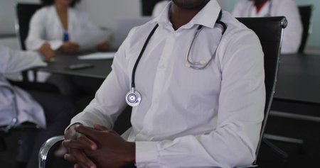 Sitting doctor in white shirt wearing stethoscope leading meeting at conference table with laptop. Professional, collaboration, healthcare, teamwork, modern, corporate, clinicalの写真素材