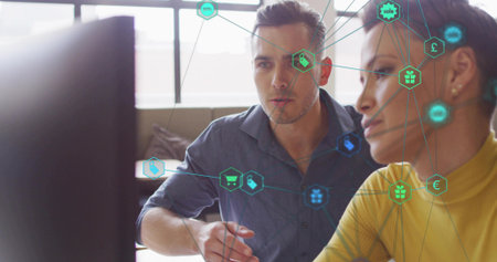 Blue-shirted man and yellow-sweatered woman reviewing e-commerce icons at office, with monitor. Technology, collaboration, innovation, digital, productivity, modern, businessの写真素材