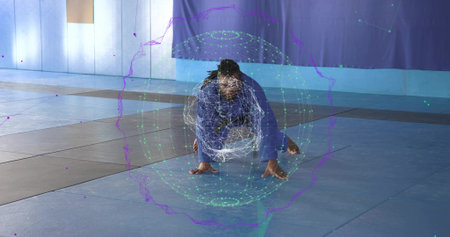Performing martial artist wearing blue gi crouching low on mat in gym, with digital hologram sphere....