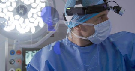 Surgeon standing in blue scrubs in operating room with surgical lamp, headlight loupe and monitor....